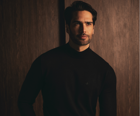 Modelo usando blusão masculino em ambiente interno com luz quente, gola simples e caimento casual, com fundo em tons amadeirados.